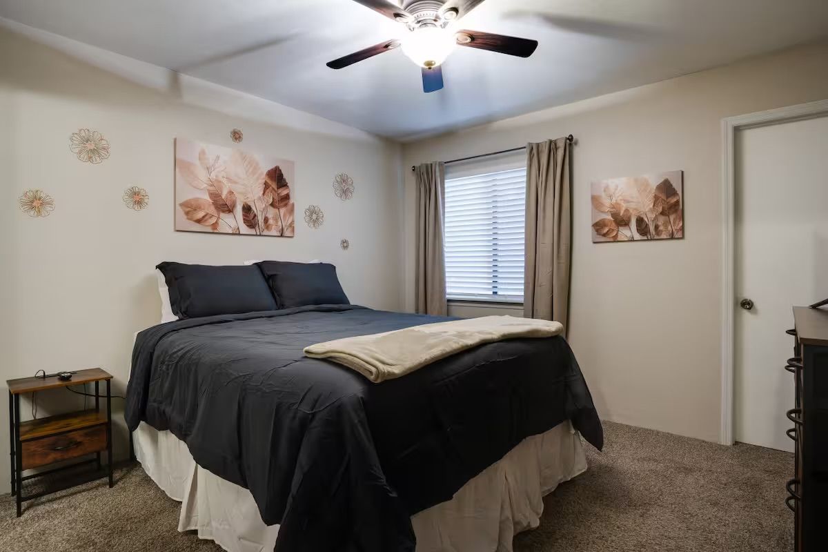 A bedroom with a bed, a ceiling fan, a window with curtains, and wall art.