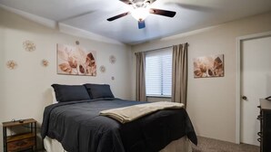 Ein Schlafzimmer mit Bett, Deckenventilator, Fenster mit Vorhängen und Wandkunst.