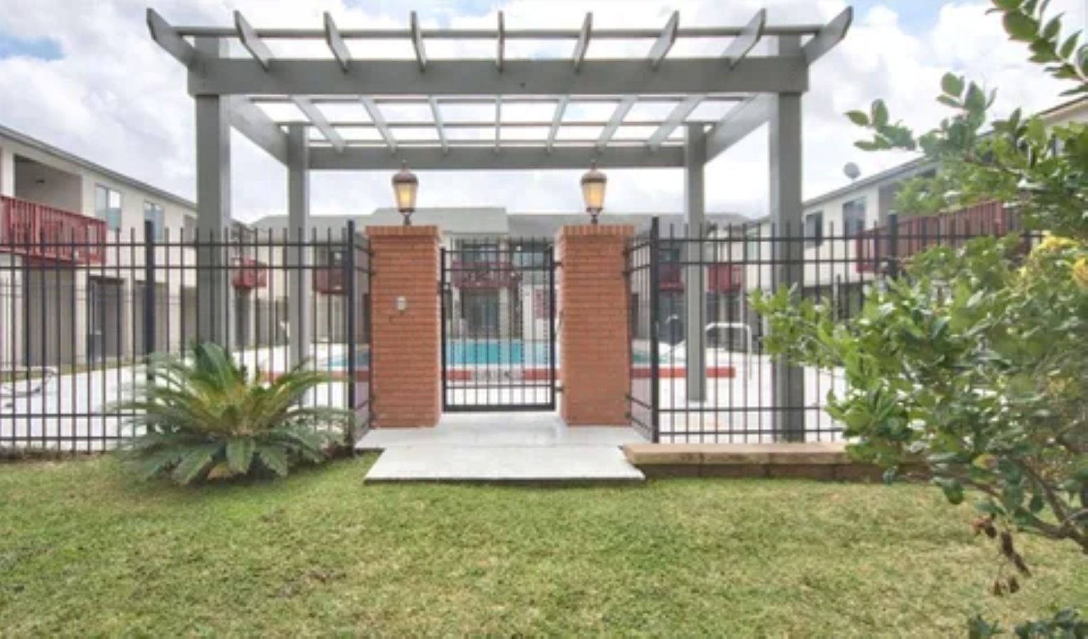 A covered outdoor area with a metal fence, brick pillars, and a pathway leading to a building entrance.
