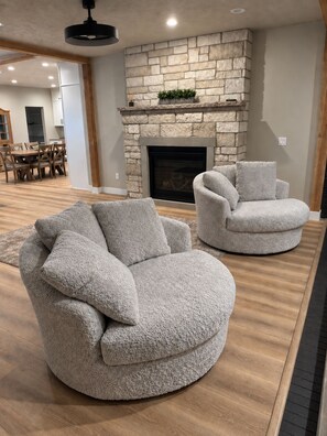A living room with a stone fireplace, two grey armchairs, and a dining area in the background.