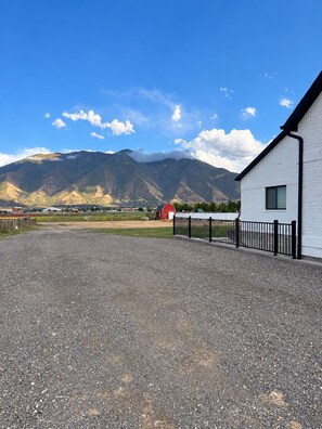 A gravel driveway leading to a white building with a black fence, a red barn in the background, and mountains under a clear sky.