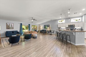 A spacious living area with a kitchen and dining space, featuring hardwood flooring, modern furniture, and large windows. - Desert Game House Private Pool, Volleyball Near Coachella (Indio)