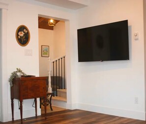 A room with a flat-screen TV mounted on the wall, a wooden floor, a staircase with a railing, and a small table with a vase. - The Old Farmhouse | Private Indoor Pool (Round Island)