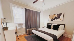 A bedroom with a bed, a radiator, a window with curtains, a ceiling fan, and a wall-mounted artwork. - Homey 1BR Space Apartment (Chicago)