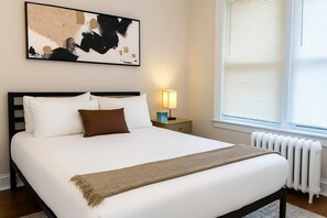 A neatly made bed with white and brown bedding, a bedside table with a lamp, and a large abstract painting on the wall. - Nice 1BR Spot, Unwind & Relax (Chicago)