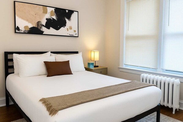 A neatly made bed with white and brown bedding, a bedside table with a lamp, and a large abstract painting on the wall.