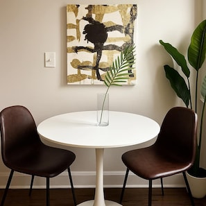 A round dining table with two chairs, a vase with a plant, a painting on the wall, and a large plant beside the table.