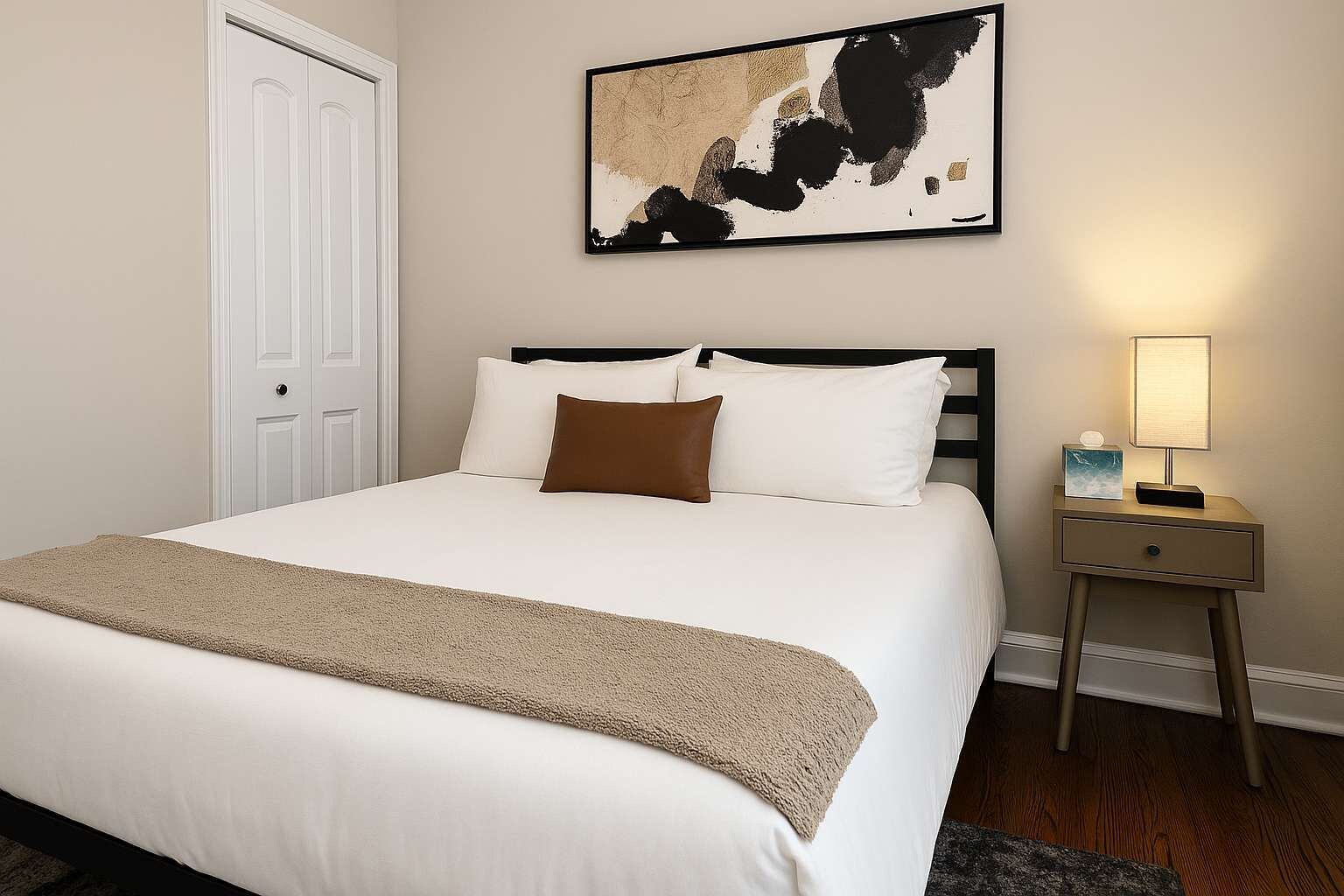 Un letto rifatto con lenzuola bianche e una coperta beige, un comodino con una lampada, e un quadro astratto appeso alla parete.
