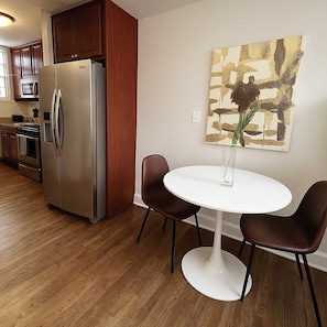 A modern kitchen with stainless steel appliances, a dining area with a white table and two chairs, and a decorative wall piece.