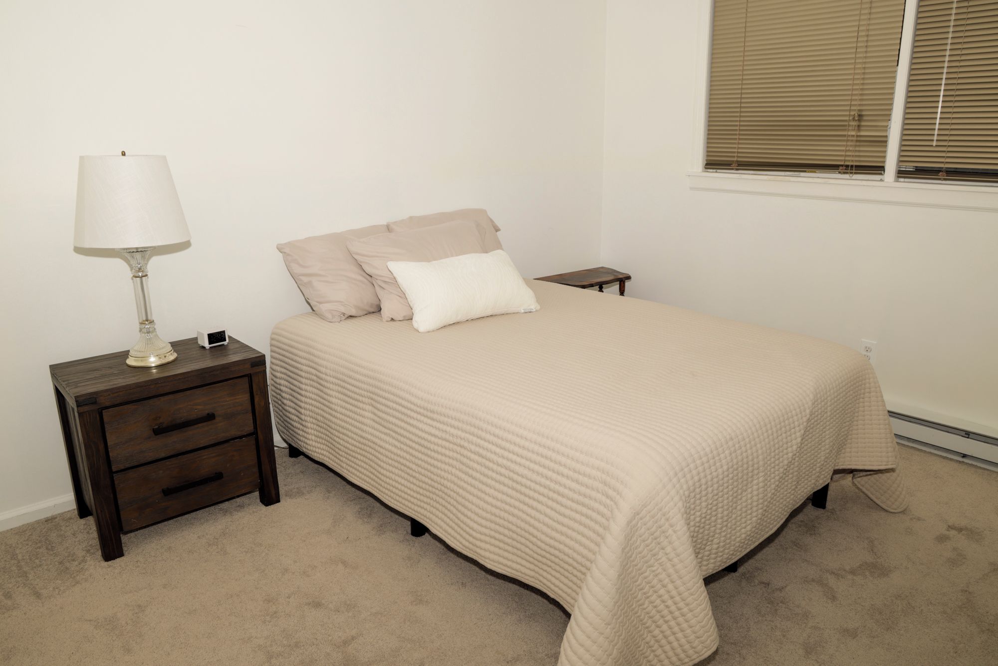 Un dormitorio con cama, mesita de noche, lámpara y ventana con persianas.