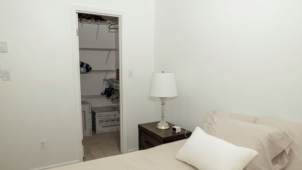 Una camera da letto con un letto, un comodino con una lampada, un armadio pieno di vestiti e una scatola etichettata 'Certified Refurbished' sul pavimento.