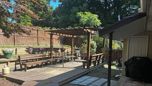 Un terrazza in legno con panche e un pergolato, circondata da un giardino con piante in vaso e un barbecue.