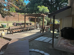 Une terrasse en bois agrémentée de bancs et d’une pergola, entourée d’un jardin avec des plantes en pot et un barbecue.