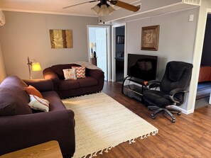 Sala de estar com sofá marrom, tapete branco, cadeira de escritório preta, televisão e ventoinha no teto.