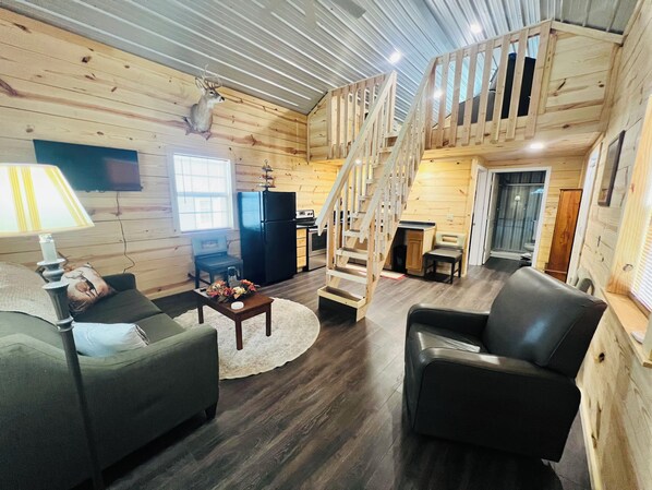 A cozy cabin interior with a living room, a kitchen, a dining area, and a staircase leading to an upper level.