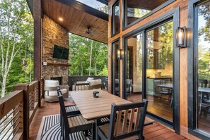 A covered outdoor deck with a stone fireplace, wooden furniture, and a glass door leading to a kitchen area. - Mountain-Modern Blue Ridge Retreat with Sauna, Hot Tub & Year-Round Views (Blue Ridge)