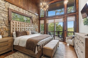 A bedroom with a stone wall, wooden ceiling, a large bed, a bench, a dresser, and a view of the outdoors. - Mountain-Modern Blue Ridge Retreat with Sauna, Hot Tub & Year-Round Views (Blue Ridge)