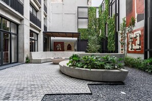 A courtyard with a brick floor, concrete planters with greenery, and a building with large windows and a mural. - Combo Suites: 4BR, Gym, Rooftop & Coworking (Mexico City)