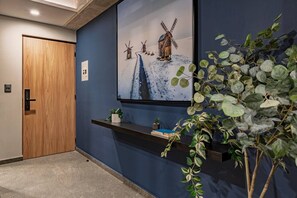 A hallway with a wooden door, a wall-mounted shelf with books and a small plant, and a painting of windmills. - Boutique 1BR Suite Near Reforma (Mexico City)