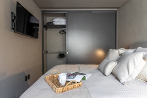 Quarto de hotel com cama, televisão de tela plana na parede, guarda-roupa e mesa de cabeceira com cesto e revista.