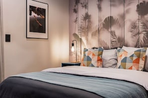 Ein Schlafzimmer mit einem Bett, Kissen, einem Nachttisch, einer Wandlampe und einem gerahmten Bild.