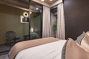 Quarto de hotel moderno com uma cama grande, área de estar com duas cadeiras, uma mesinha e uma planta em vaso.