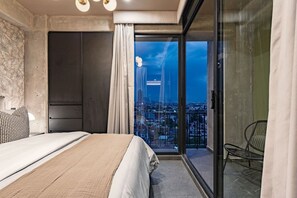 Ein Schlafzimmer mit einem großen Bett, Blick auf die Stadt durch eine Glastür und ein Balkon mit einem Stuhl.