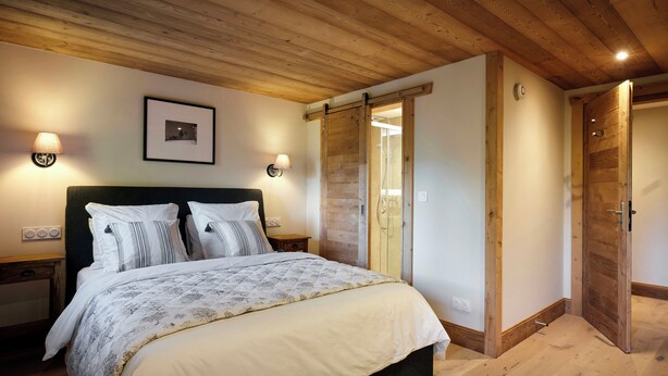 Quarto com teto de madeira, cama com roupa de cama branca, duas luminárias de cabeceira e uma porta que dá para uma área de chuveiro.