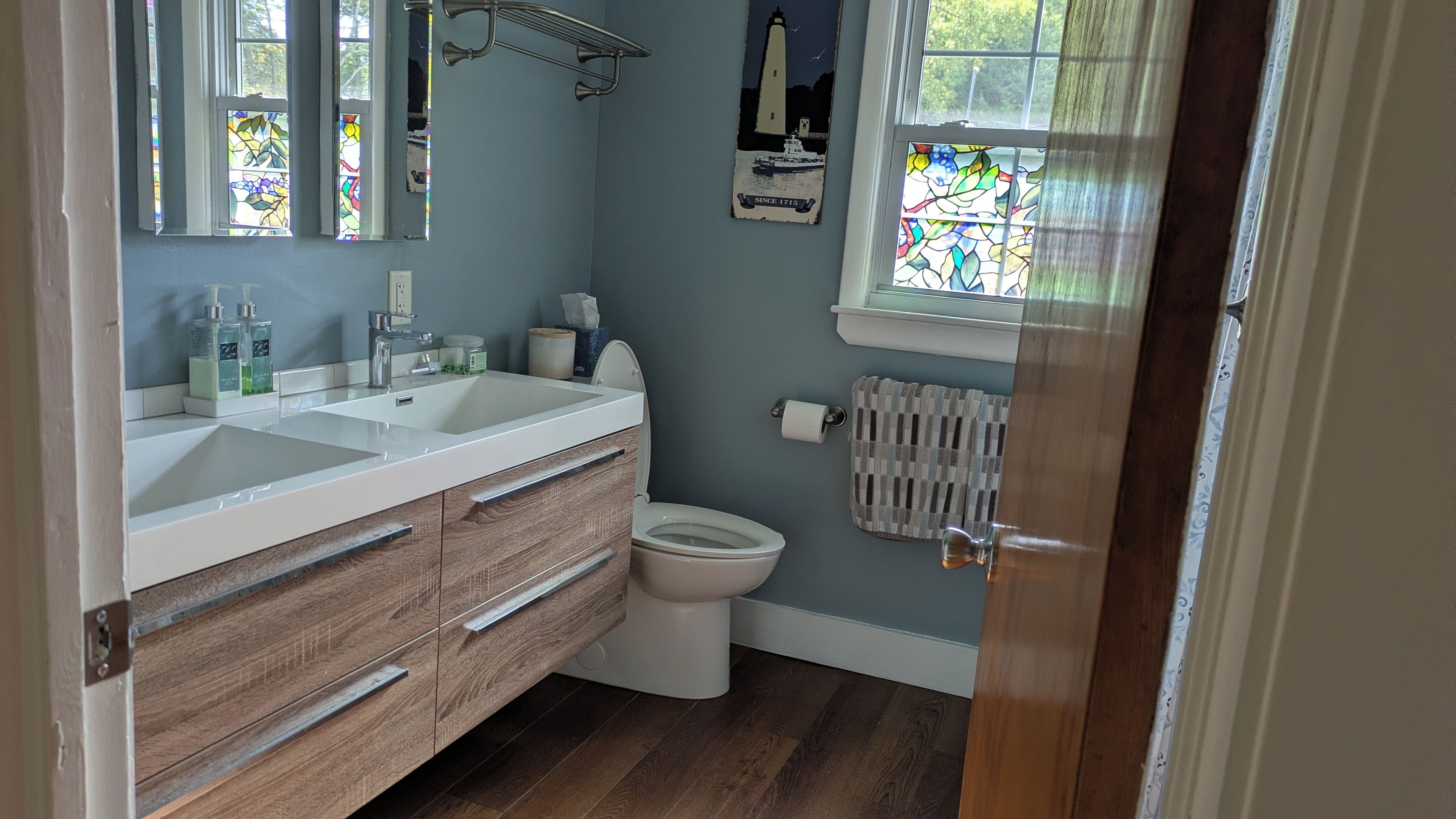 Un baño con lavamanos doble, tocador de madera, inodoro y ventana con vistas a un faro.