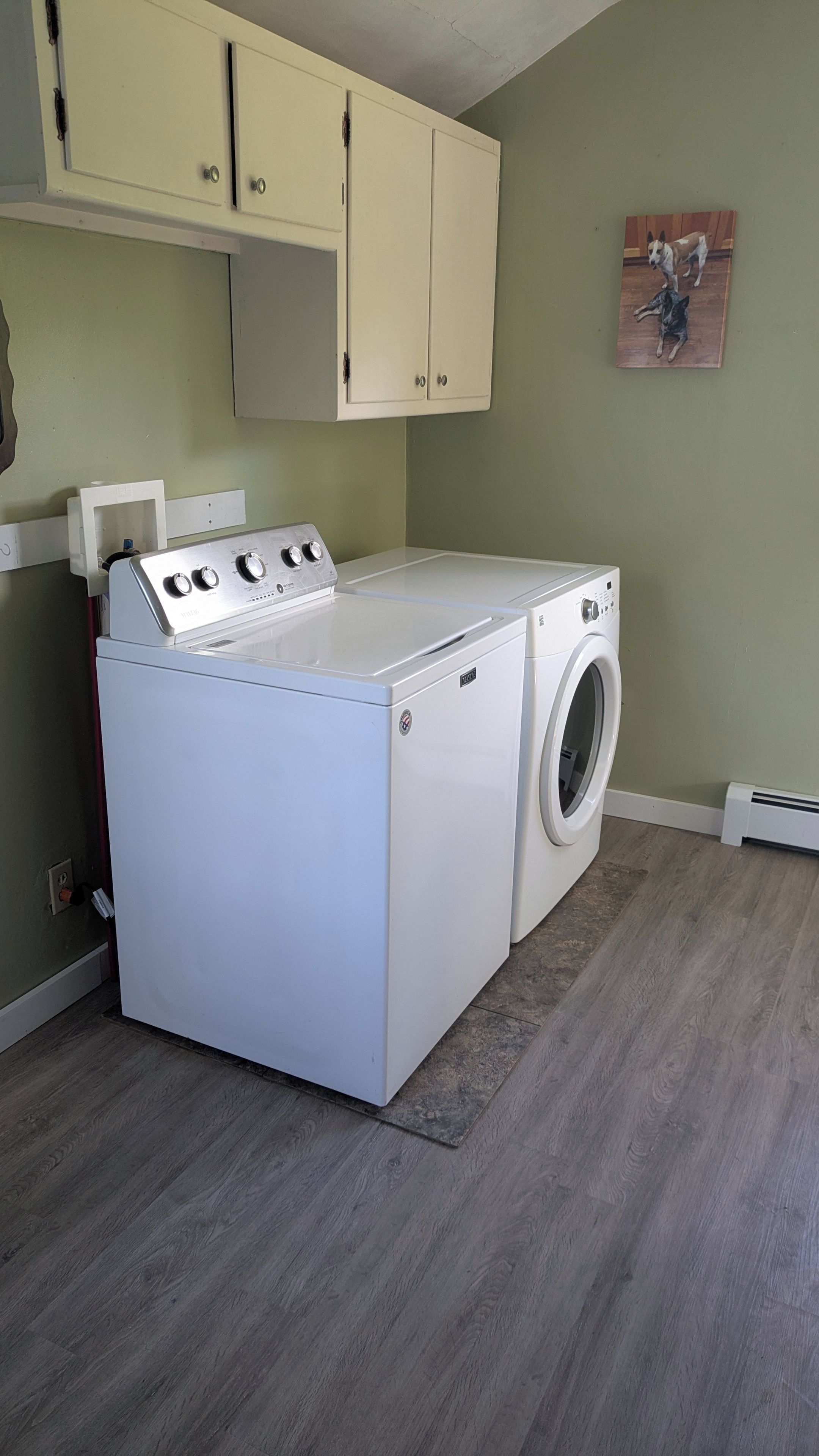 A laundry room with a front-loading washer and dryer, wooden cabinets above, and a painting on the wall.
