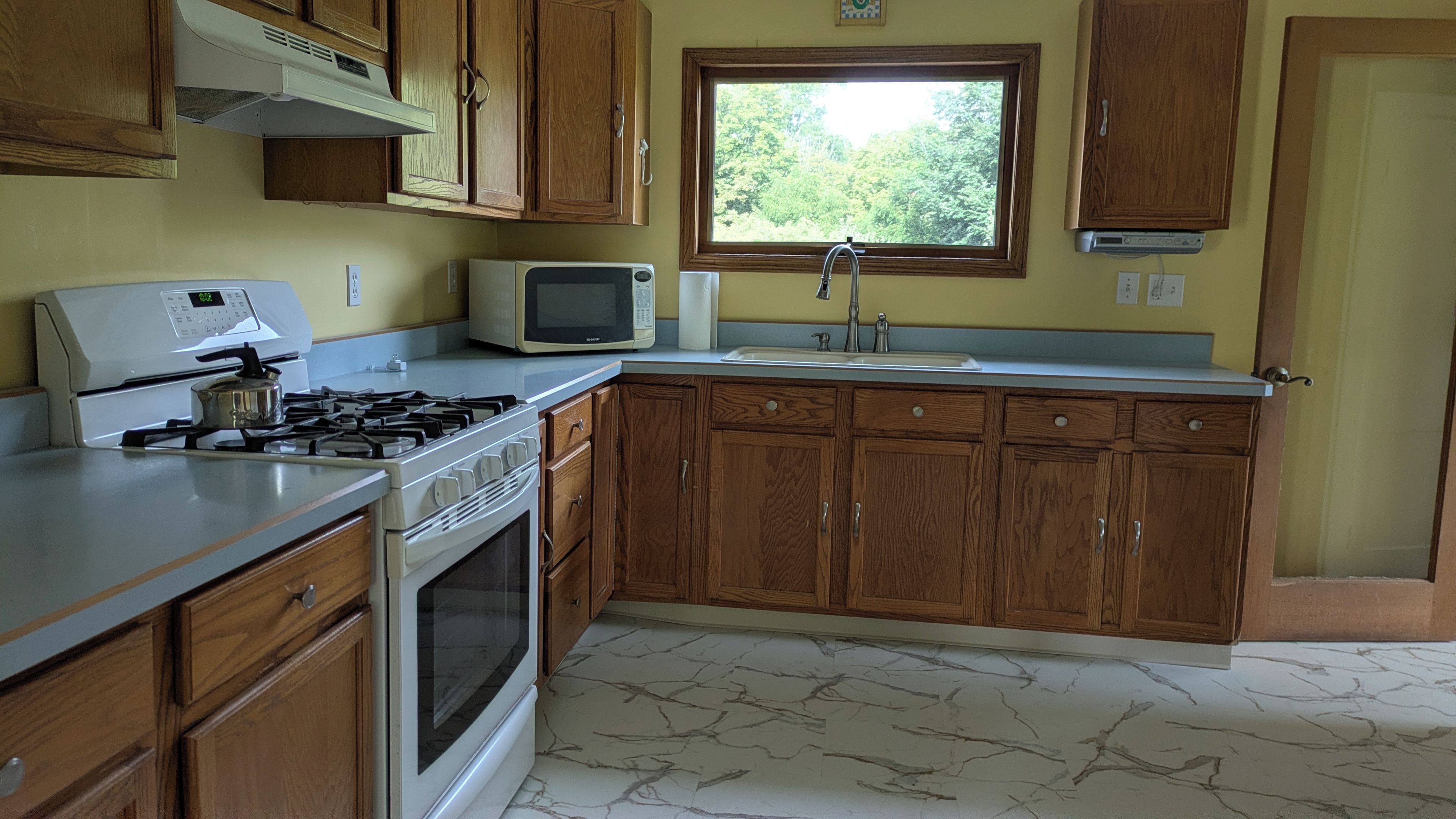 A kitchen with wooden cabinets, a gas stove, a microwave, and a window with a view of greenery.