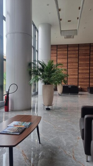 En rummelig lobby med marmorgulv, en receptionsdisk, en stor potteplante og et moderne receptionsområde med en væg beklædt med træpaneler.