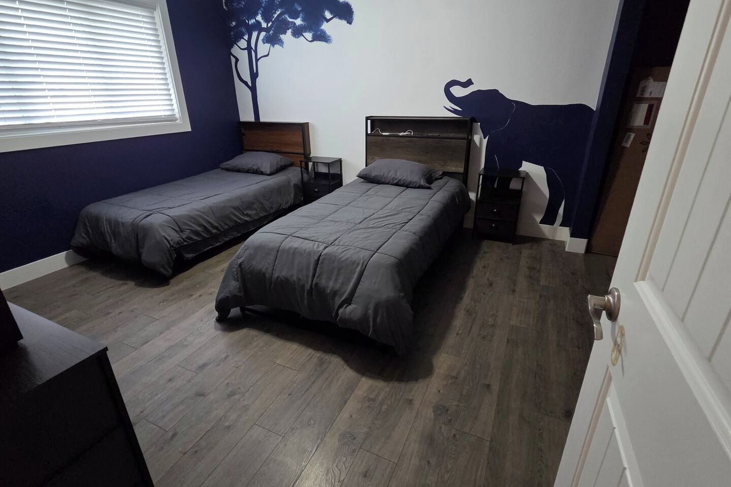 A bedroom with two single beds, a nightstand, a dresser, a window with blinds, and a wall decal of a tree and a cow.