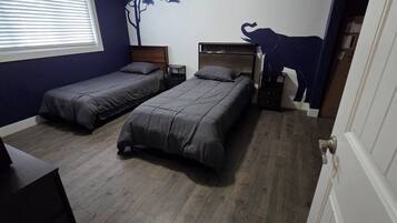 A bedroom with two single beds, a nightstand, a dresser, a window with blinds, and a wall decal of a tree and a cow.