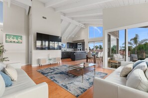 Un salon moderne avec une table basse en verre, des canapés blancs et une vue sur la cuisine et la terrasse extérieure.