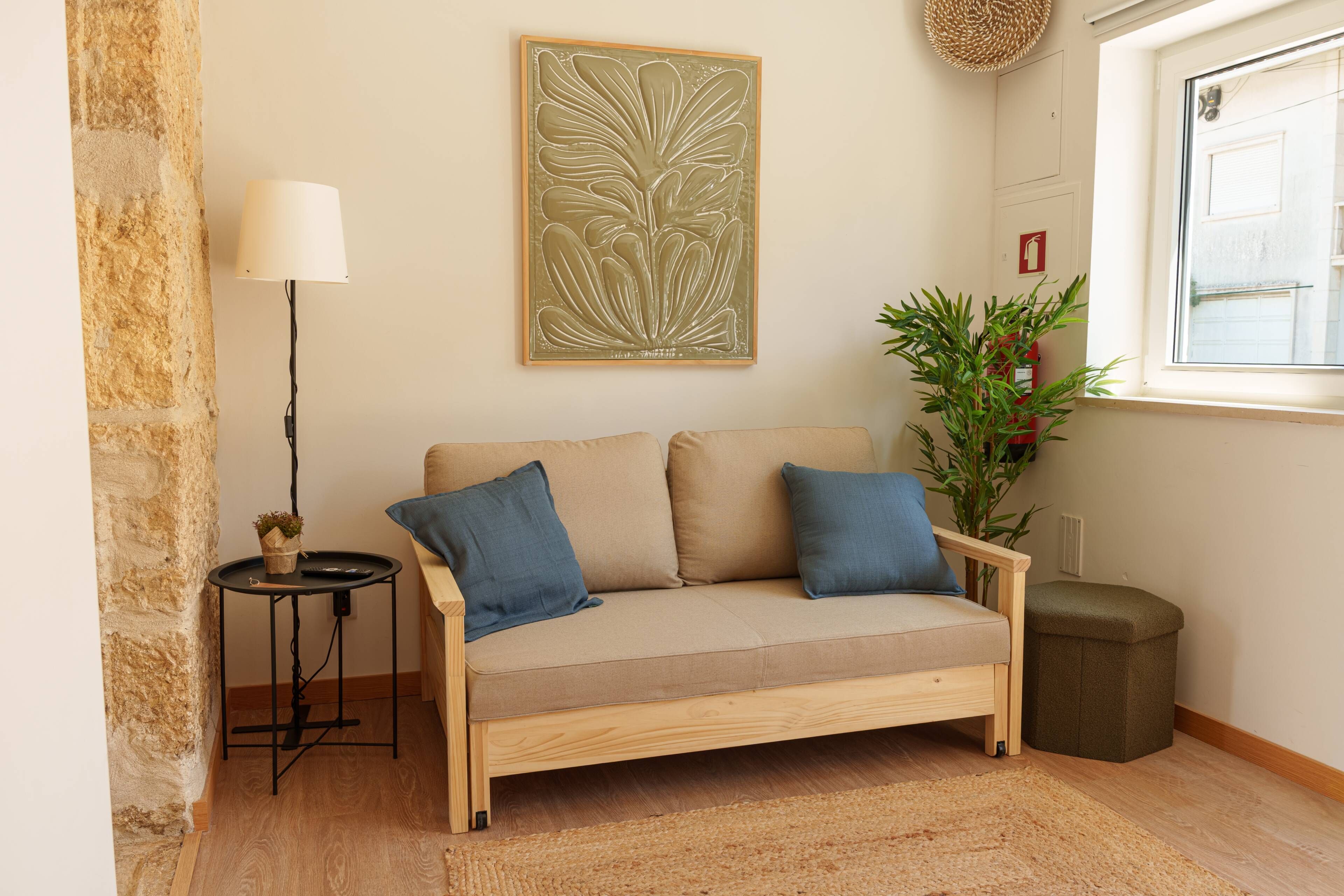 Ein Zimmer mit einem beigen Sofa, zwei blauen Kissen, einem kleinen Beistelltisch mit einer Lampe, einer Topfpflanze und einem Feuerlöscher.