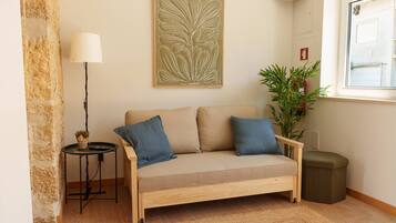 Ein Zimmer mit einem beigen Sofa, zwei blauen Kissen, einem kleinen Beistelltisch mit einer Lampe, einer Topfpflanze und einem Feuerlöscher.