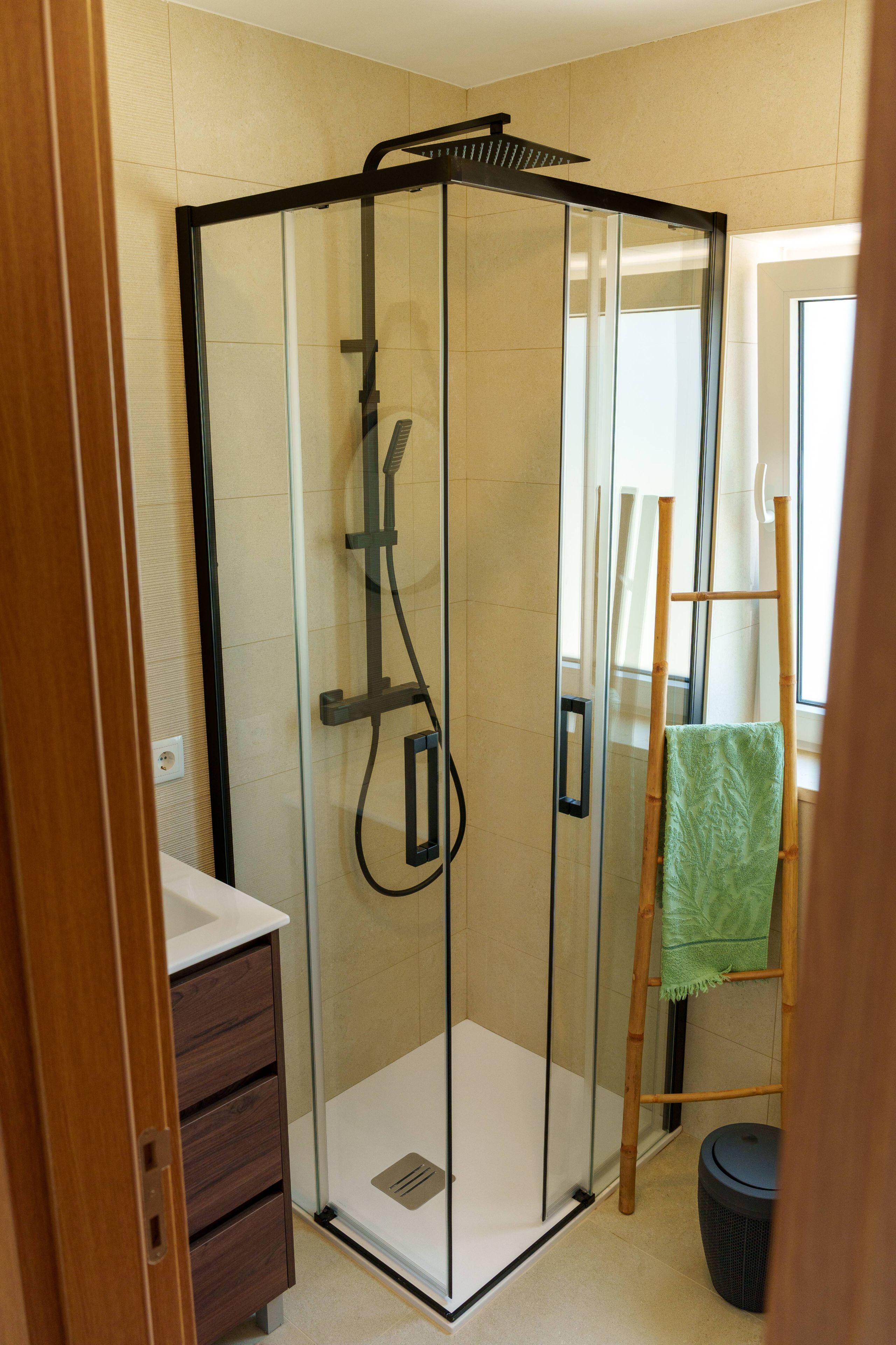 A glass-enclosed shower with a control panel, a wooden cabinet, and a green towel on a wooden chair.