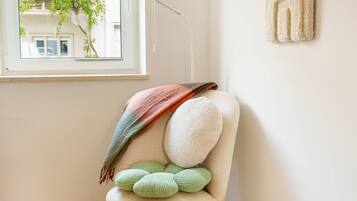 Una habitación con ventana, una silla con cojines, un tapete redondo y una obra de arte en la pared.