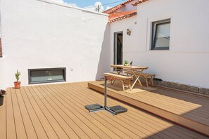 Una terraza de madera con una mesa, sillas, una planta en maceta y una ventana.