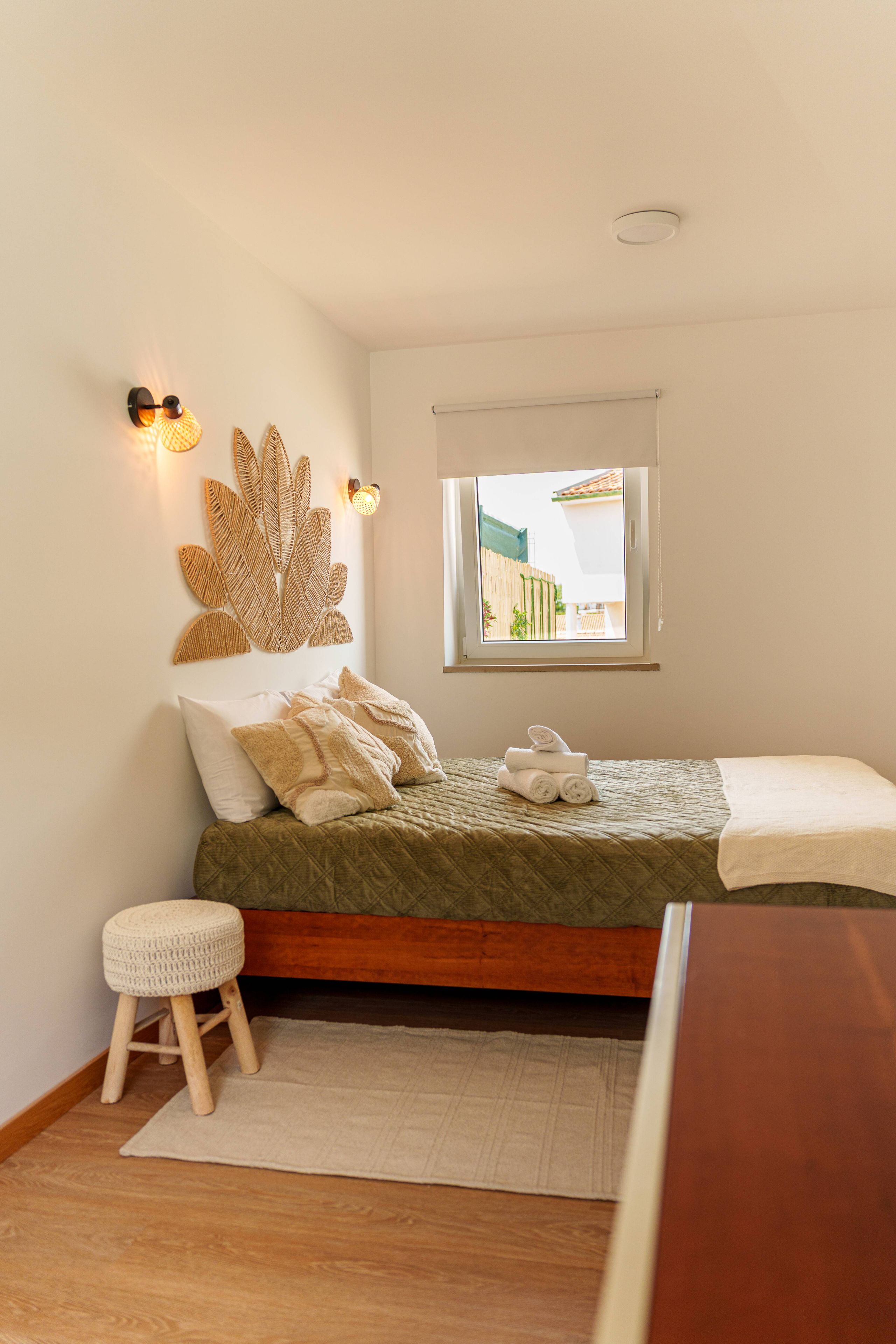 A bedroom with a bed, a window, a wall-mounted light fixture, and a small stool.
