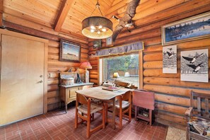 A rustic cabin interior with a dining table, chairs, a lamp, a fireplace, and framed artwork on the walls. - Cozy Riverfront Log Cabin with Private Hot Tub (Bend)