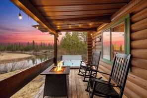 A wooden cabin with a fire pit, two chairs, and a hot tub on a wooden deck overlooking a river. - Cozy Riverfront Log Cabin with Private Hot Tub (Bend)