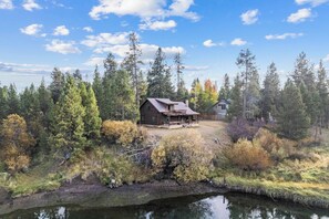 A rustic cabin with a porch, surrounded by dense forest and a small body of water. - Cozy Riverfront Log Cabin with Private Hot Tub (Bend)