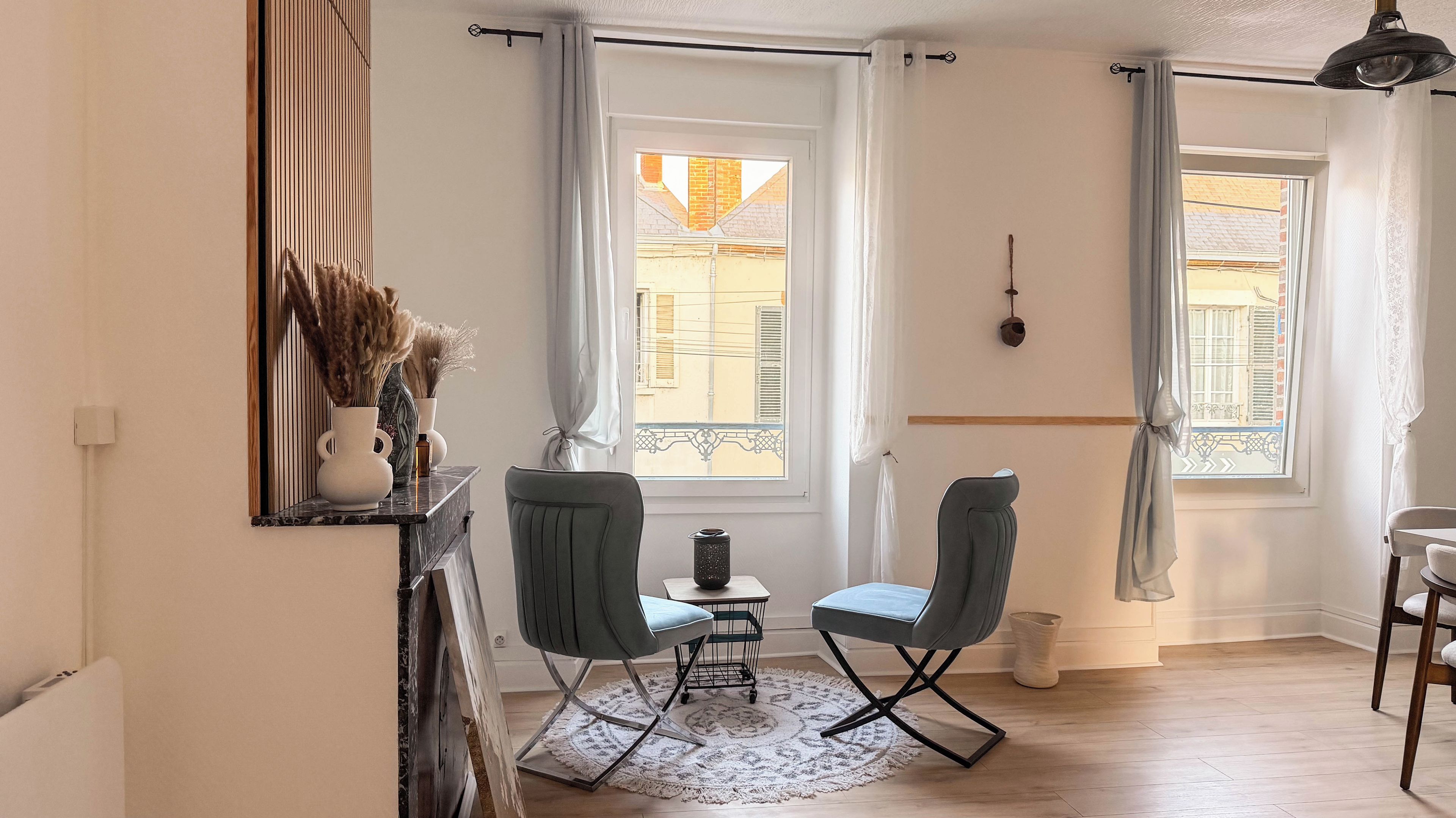 Ett rum med två stolar, ett litet bord, en vas med torkade växter och ett fönster med utsikt över byggnader.