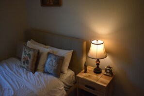 Ein ordentlich bezogenes Bett mit Kissen, einer Nachttischlampe und einer kleinen Uhr auf einem Holznachttisch.