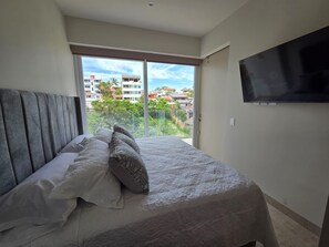 A bedroom with a bed, a TV on the wall, and a balcony with a view of buildings. - Vrbo Property (Bucerías)