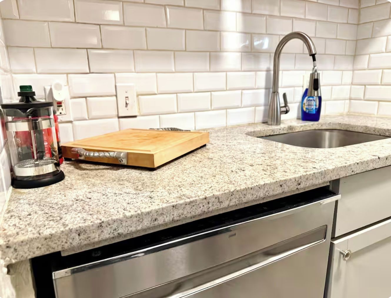 A kitchen with a granite countertop, a wooden cutting board, a stainless steel sink, and a dishwasher.