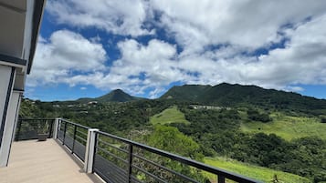 A balcony with a metal railing overlooking a mountainous landscape with greenery and a clear sky.