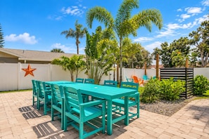Une terrasse extérieure avec une table et des chaises turquoise, entourée de palmiers et d’une clôture blanche.